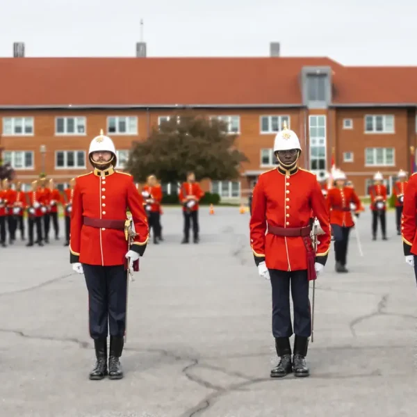 A group of people in red military uniforms and white helmets stand in formation on a paved courtyard, with a brick building and trees in the background.
