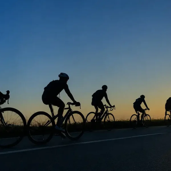 Six cyclists ride in a line along a road at sunset, their silhouettes dark against the orange and blue sky. The sun is low on the horizon, casting long shadows as they pedal together.