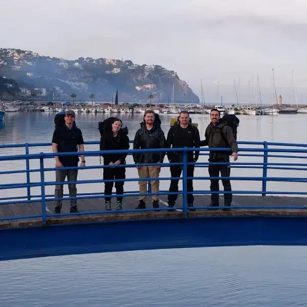 Five people with backpacks stand on a blue footbridge over water, with sailboats and a hilly shoreline in the background under a cloudy sky.