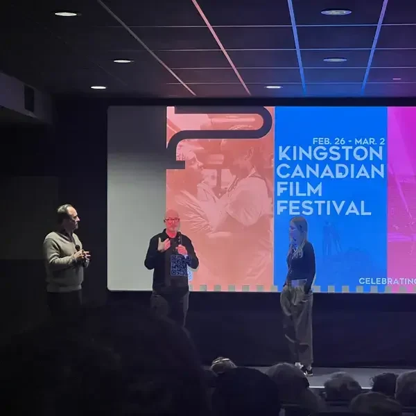 Three people stand on a stage in front of a screen displaying “Kingston Canadian Film Festival” with dates. The audience is visible in the foreground, and the theater is dimly lit.