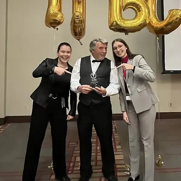 Three people pose smiling at an indoor event, with two women pointing at a man in a tuxedo who is holding an award. Large gold balloons form letters behind them.