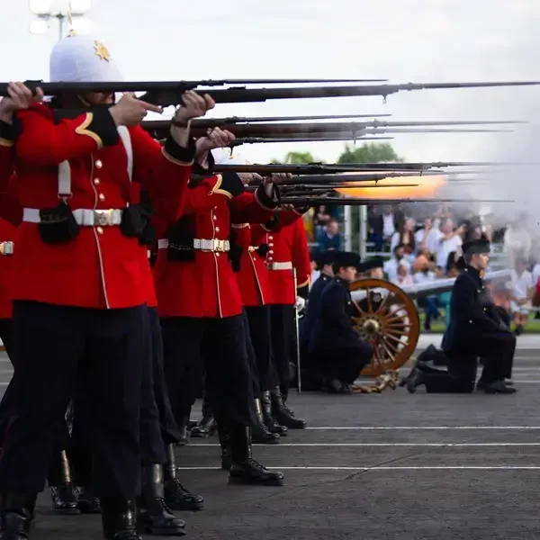 Des soldats en uniforme rouge tirent à l'arme à feu en formant un salut synchronisé, tandis qu'à l'arrière-plan, des hommes en uniforme sombre actionnent un canon, d'où s'échappe de la fumée. Des spectateurs assistent à la scène depuis les tribunes.