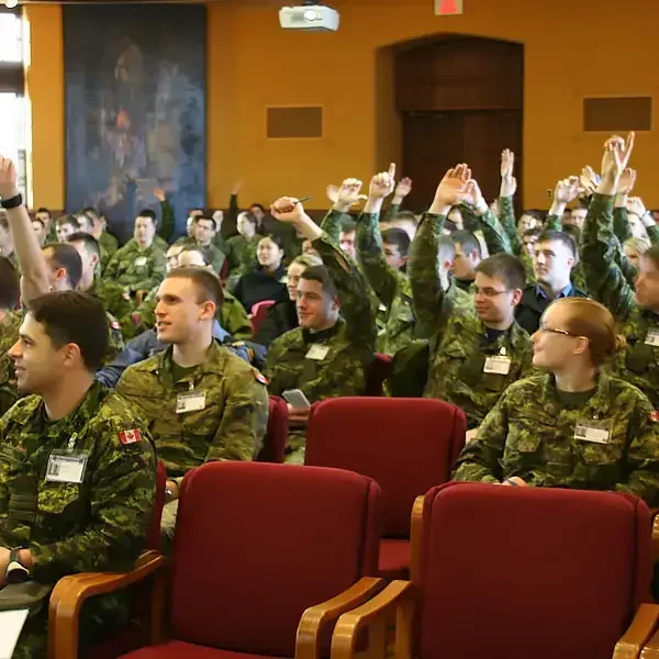 Un groupe de personnes vêtues d'uniformes de camouflage est assis sur des chaises rouges ; beaucoup lèvent la main, donnant l'impression de participer à un cours ou à un séminaire.