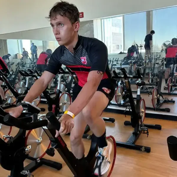 A young person in athletic gear rides a stationary bike in a gym with mirrors, focused on their workout. Several other people are also cycling in the background.