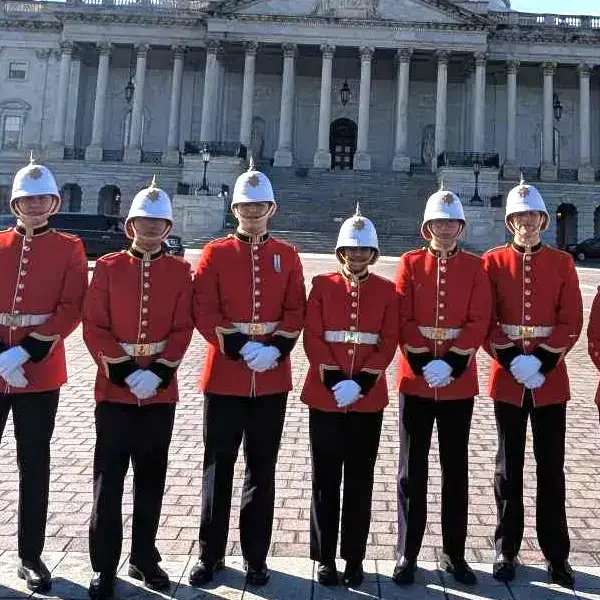 Seven people in red British-style ceremonial uniforms and white helmets stand in a line outside a large, columned building on a sunny day.