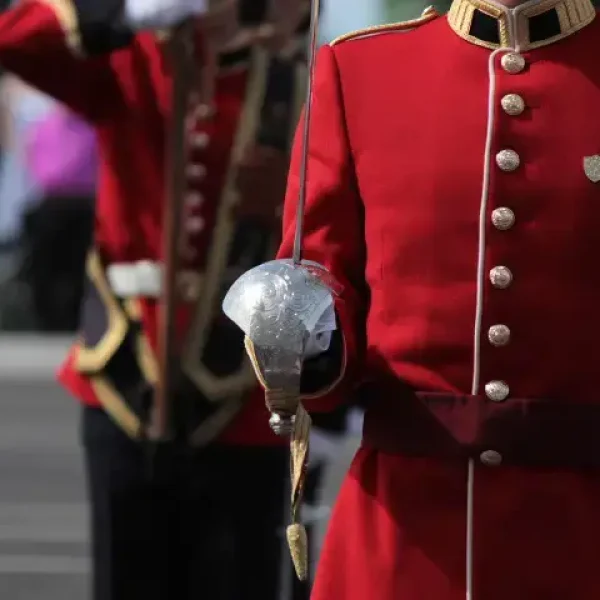 A person in a red military uniform holds a sword with a curved guard. Another similarly dressed figure stands in the background, saluting. The scene suggests a formal or ceremonial event outdoors.