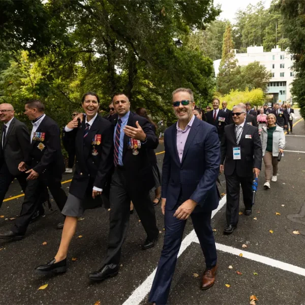 A group of people in formal attire, some with medals, walk outdoors on a wooded path. Some are smiling and talking, while others walk behind them. Trees and a white building are visible in the background.