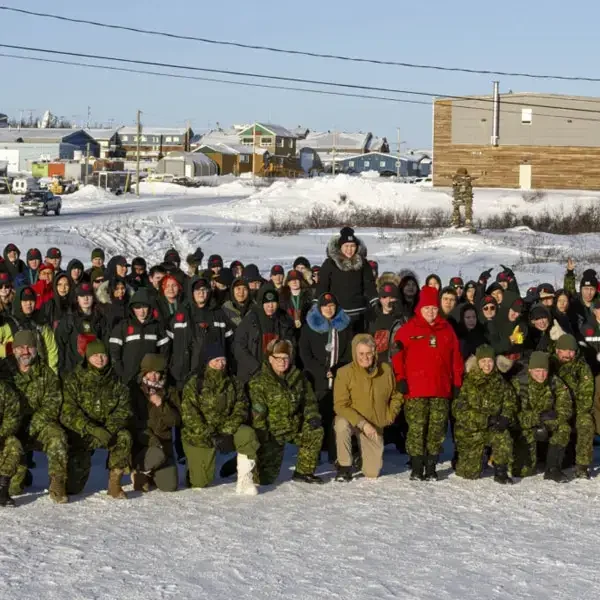 A large group of people in winter clothing, including camouflage uniforms, pose for a group photo on snowy ground with buildings and vehicles in the background.