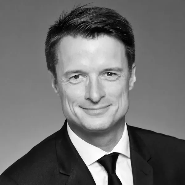 Smiling man in a suit and tie, with short dark hair, against a plain gradient background. The photo is black and white and appears to be a professional headshot.