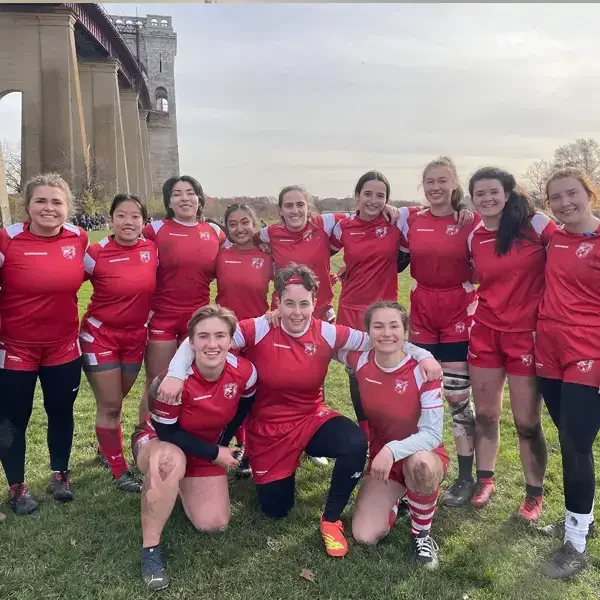 A women’s rugby team in red uniforms poses together outdoors on grass, smiling, with an arched bridge structure in the background.