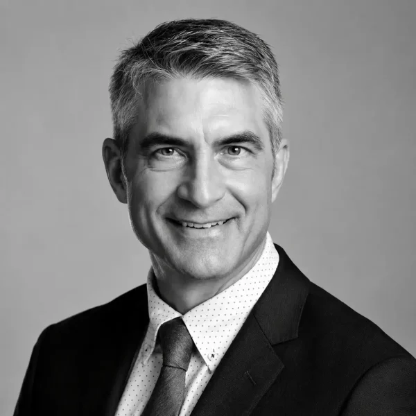 Smiling middle-aged man with short gray hair wearing a dark suit, tie, and light polka-dot shirt, posing in front of a plain background. The image is in black and white.