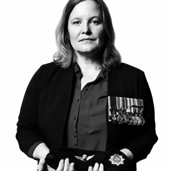 A woman with medals on her jacket holds a beret and a military badge in her hands, facing the camera with a serious expression against a plain white background.