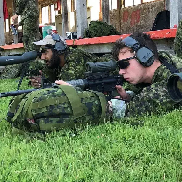 Deux soldats en tenue de camouflage et équipés d'écouteurs sont allongés dans l'herbe, leurs fusils pointés vers une cible. L'un d'eux utilise une lunette de visée et tous deux affichent un air grave, concentrés sur leur cible. On aperçoit un sac à dos de camouflage et un bâtiment à l'arrière-plan.