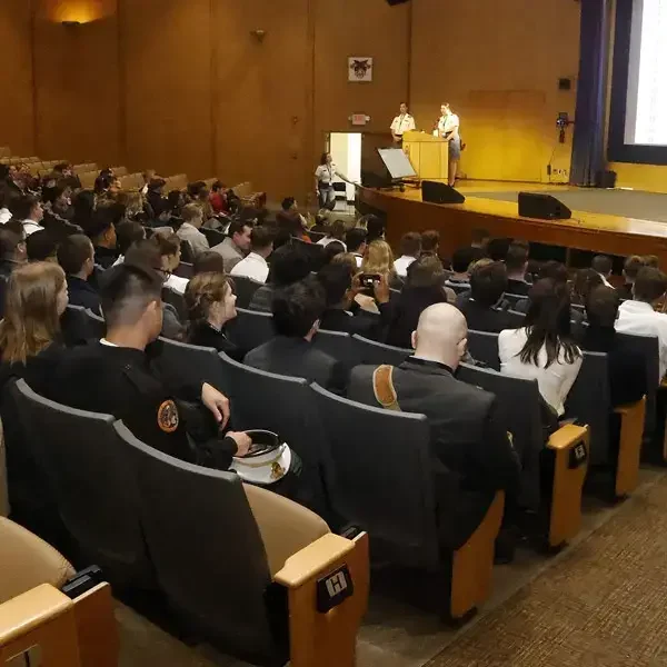 Des personnes en tenue professionnelle sont assises dans un auditorium et regardent deux intervenants sur scène. Un grand écran et un podium sont visibles au fond de la salle.